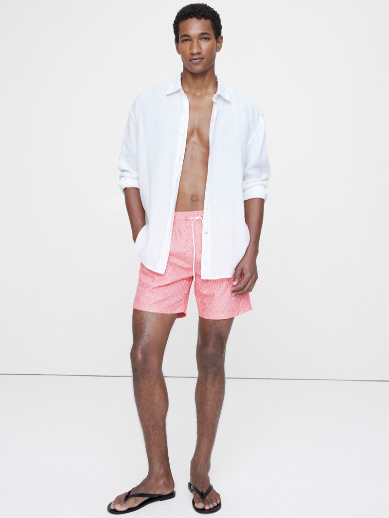 Model wears white linen shirt open with salmon color swim shorts and black flip flops