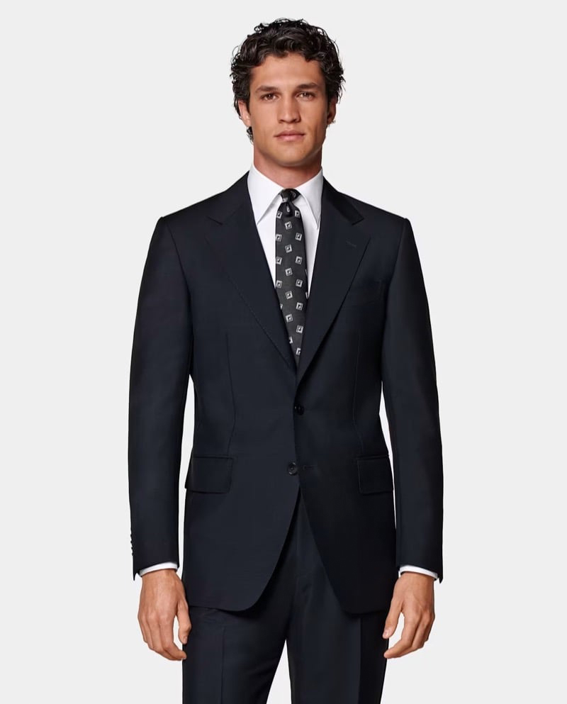 Model wears a dark navy single-breasted suit with a white shirt and geometric print tie