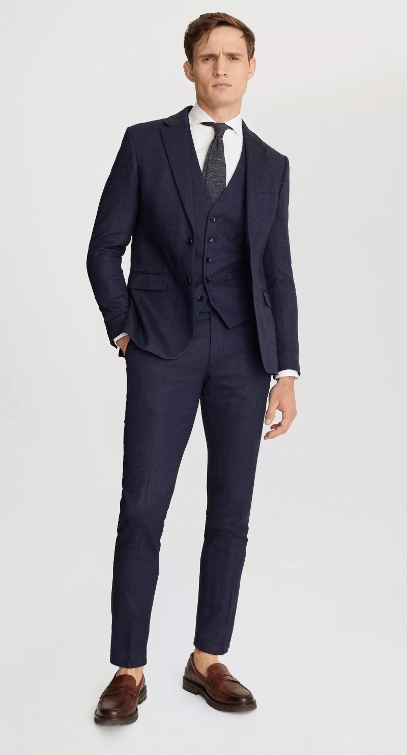 Model wears a three-piece navy suit with a white dress shirt, charcoal tie, and brown leather loafers