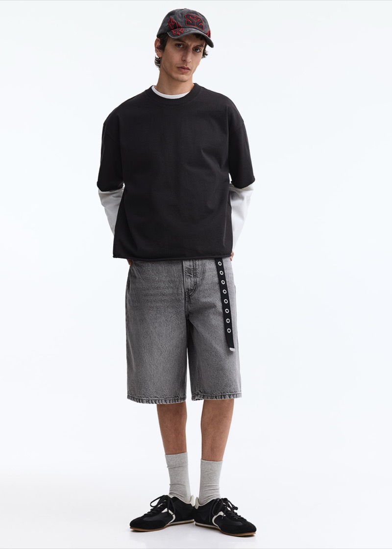 Man wearing baggy grey denim shorts with layered tees and a cap