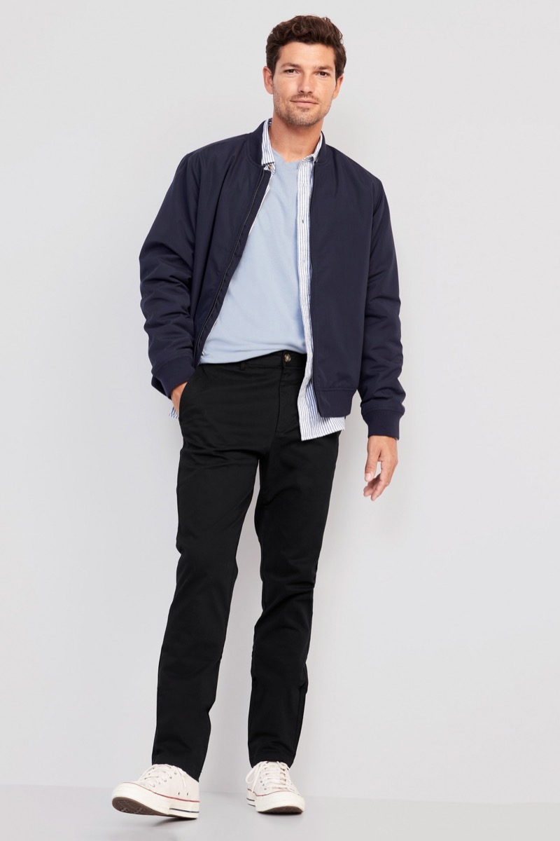 Man wearing slim black chinos with navy bomber jacket open over blue striped button-down and light blue tee with white Converse sneakers