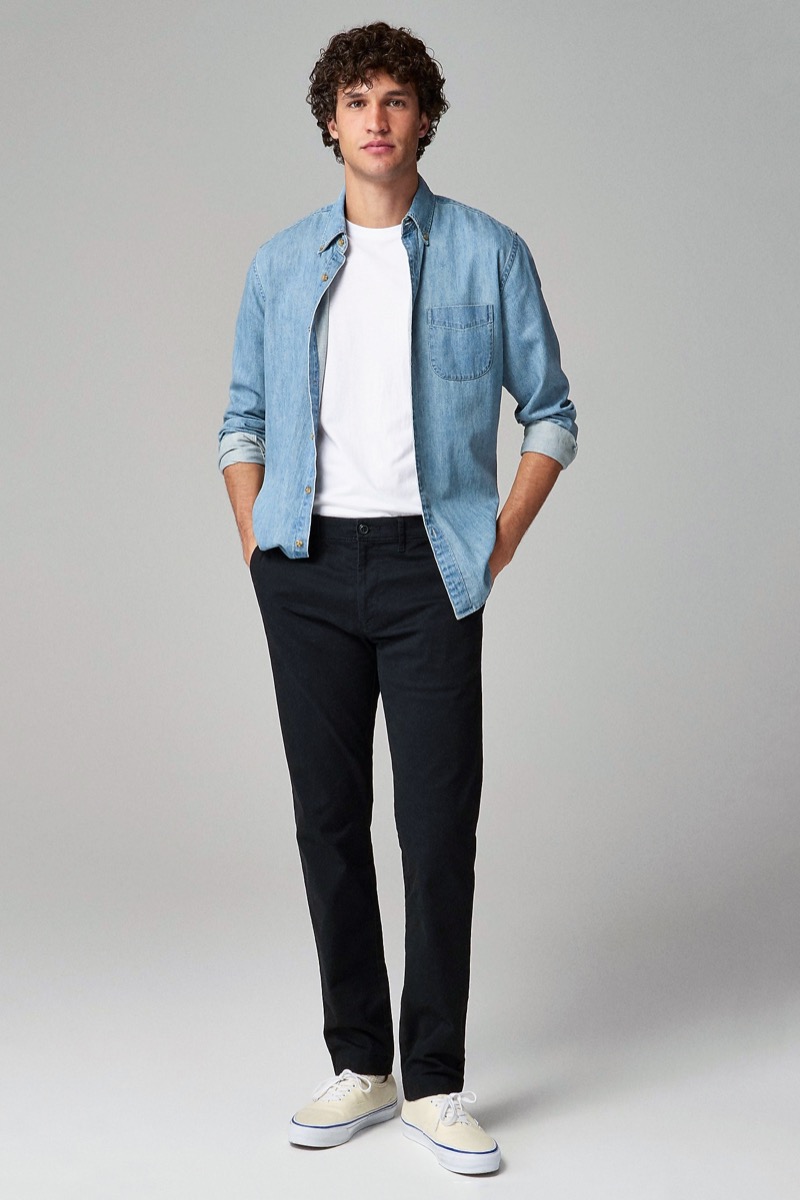 Man wearing slim black chinos with light wash denim shirt open over white tee and off-white canvas sneakers