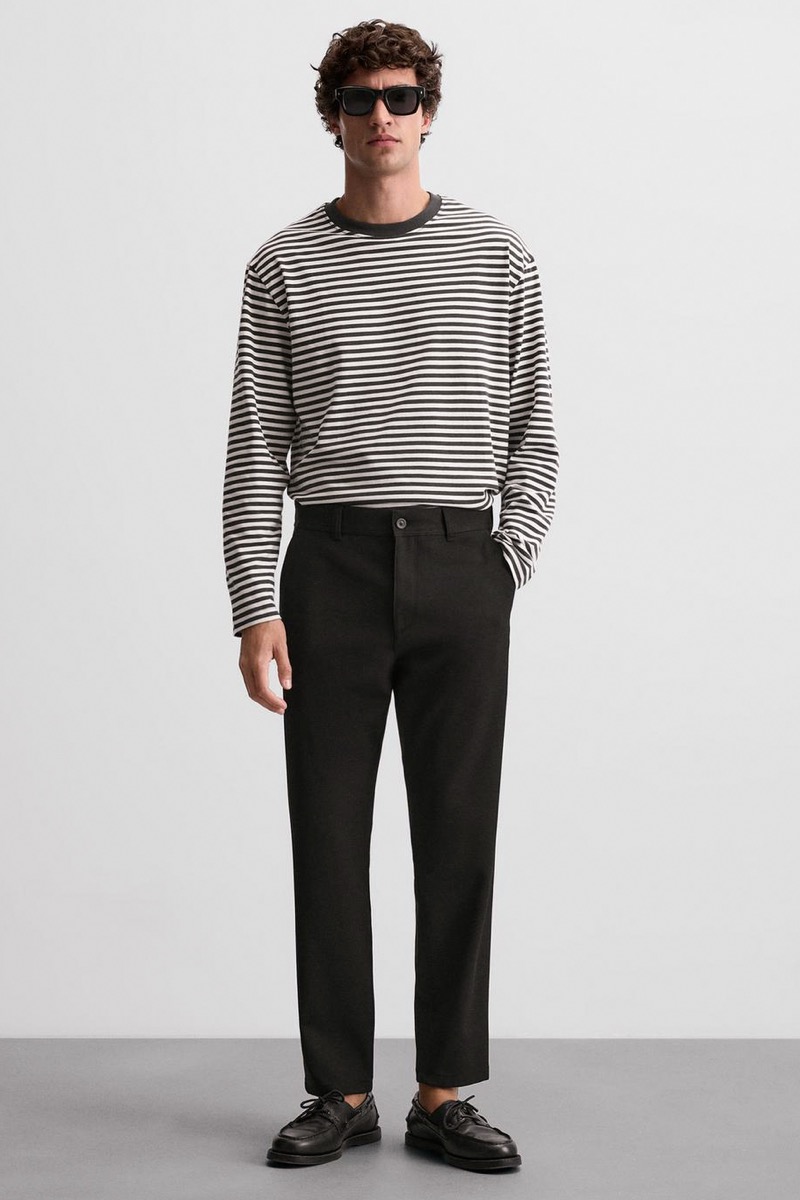 Man wearing tapered black chinos with oversized black-and-white striped long-sleeve tee, sunglasses, and black loafers
