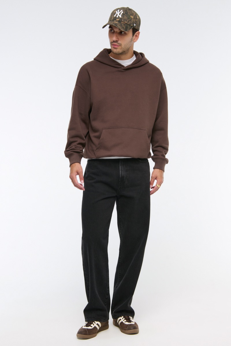 Man wearing wide-leg black jeans with brown oversized hoodie over white tee, camo NY baseball cap, and brown-and-white terrace sneakers