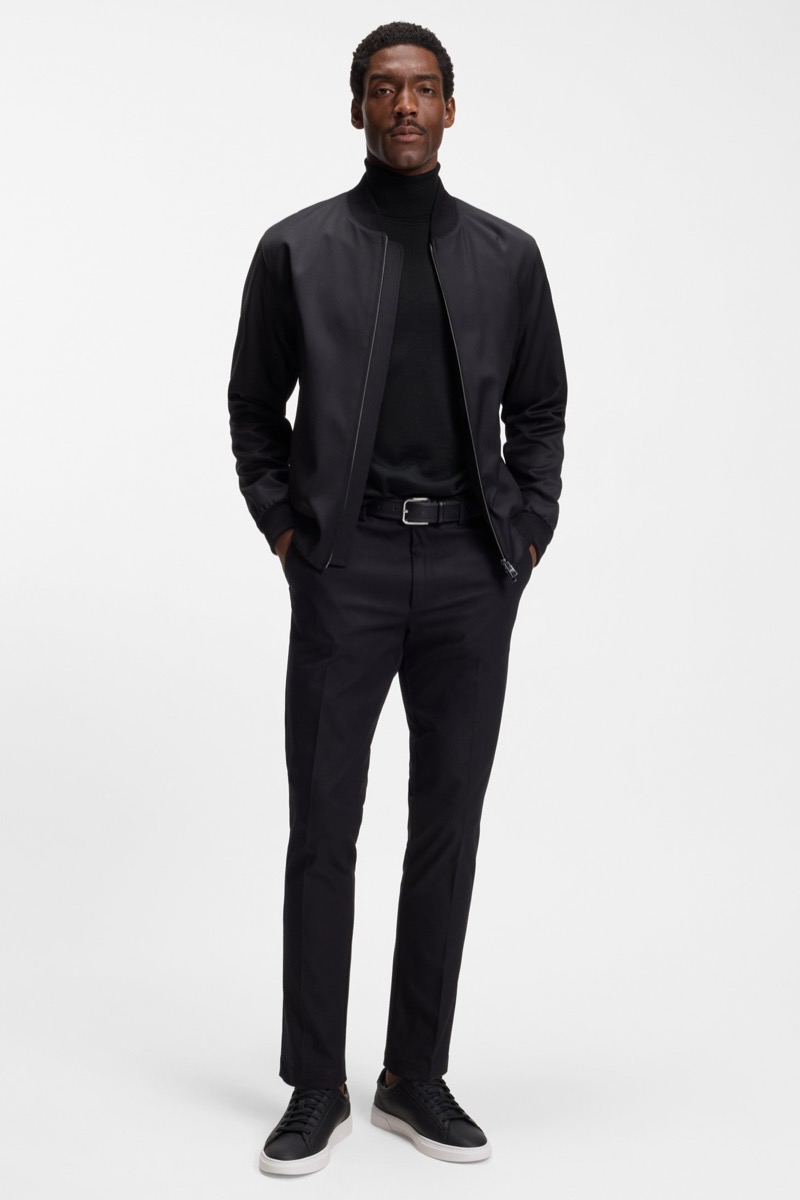 Man wearing slim black dress pants with black turtleneck, black tech jacket, black leather belt, and black leather sneakers