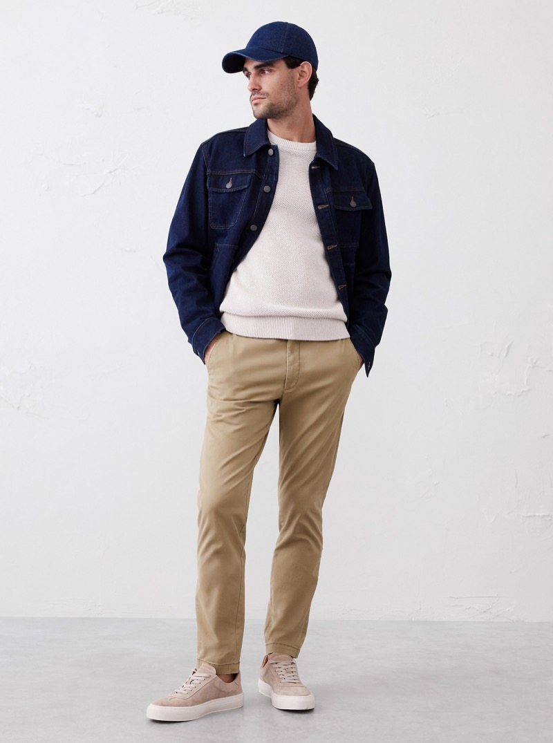 model wears khaki chinos with a denim jacket, crew sweater, cap, and sneakers