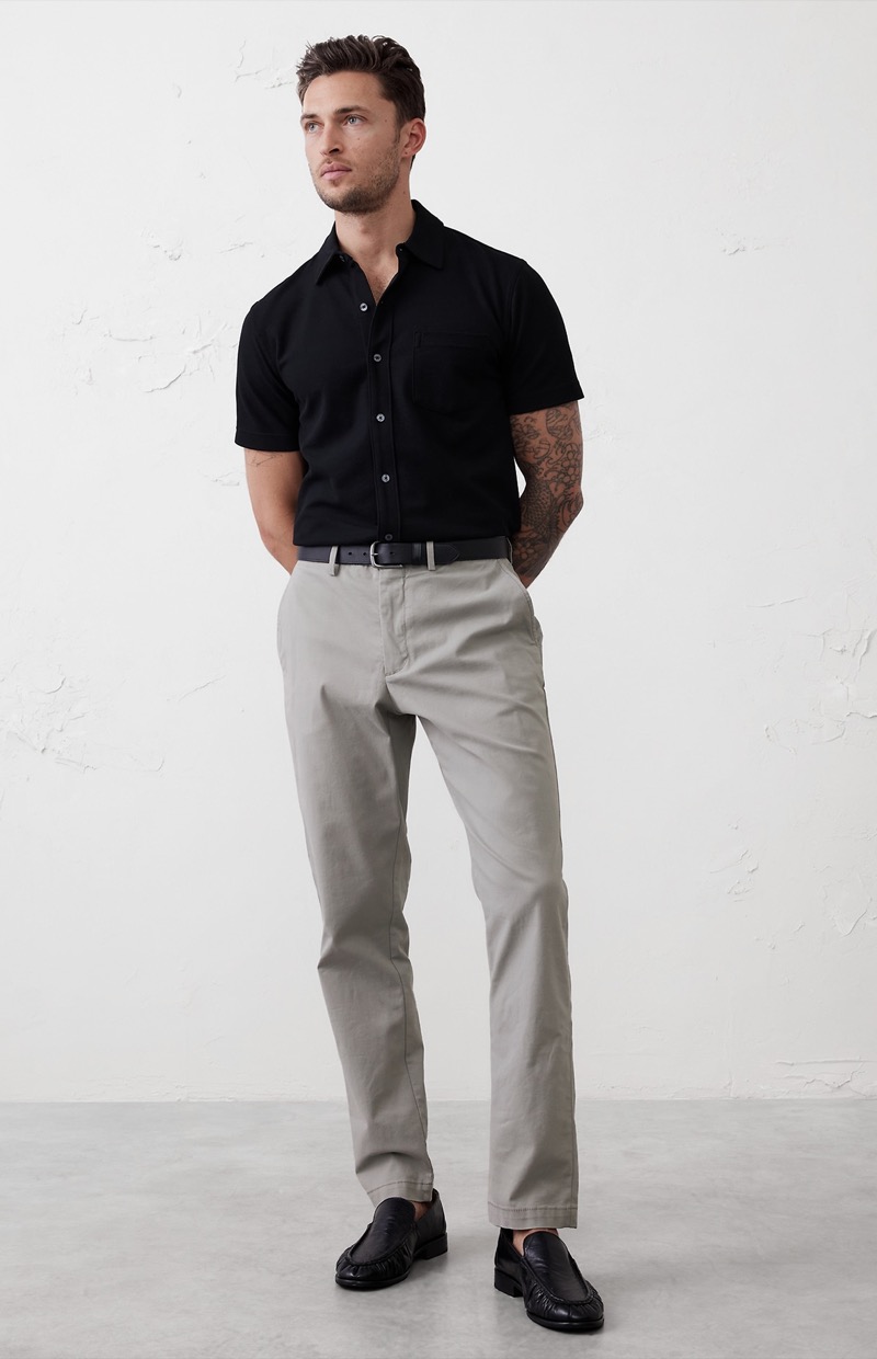 model wears gray chinos with black short-sleeve button-down shirt and black leather loafers