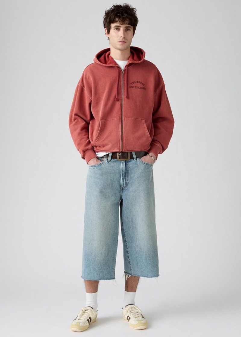 Man wearing long light wash denim shorts with a red cropped zip hoodie and vintage sneakers