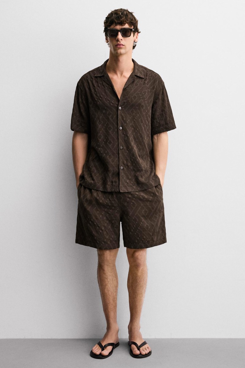 Man wearing a dark brown textured camp collar shirt with matching dark brown textured shorts, sunglasses, and black leather thong flip-flops