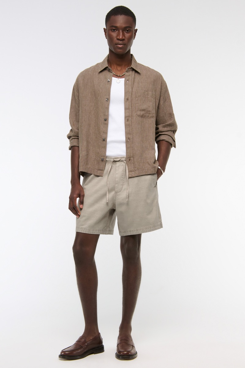 Man wearing a tan pinstripe linen overshirt open over a white tank top with stone drawstring shorts and dark brown leather penny loafers