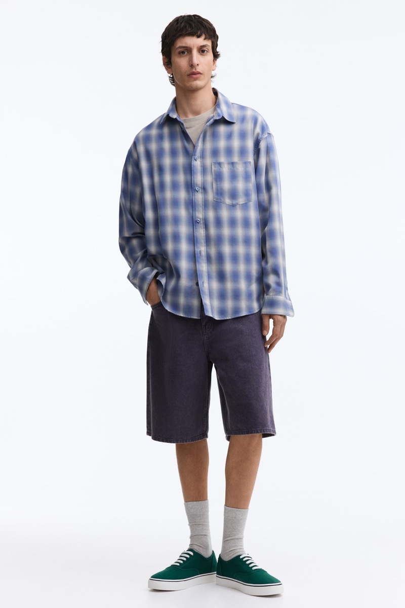 Man wearing an oversized blue and white plaid flannel shirt with washed baggy dark denim shorts, grey crew socks, and dark green canvas low-top skate sneakers