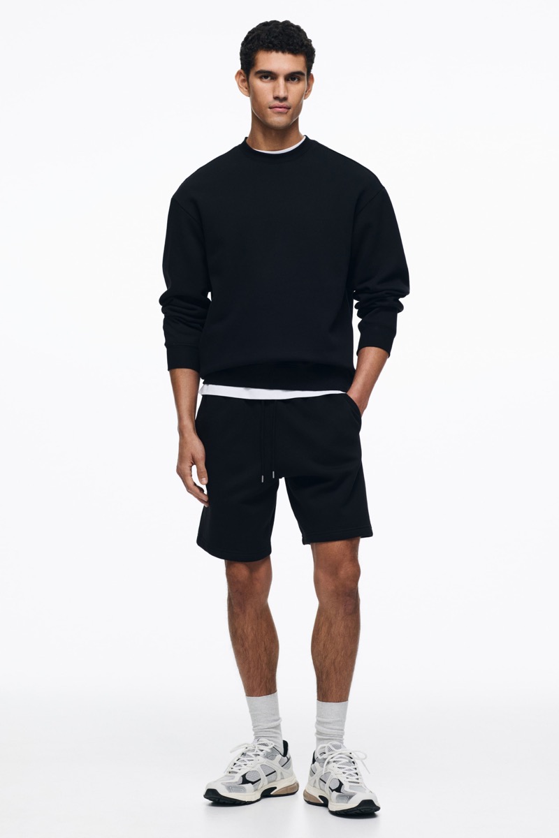 Man wearing a black crewneck sweatshirt, black fleece shorts, grey crew socks, and chunky white and grey running sneakers