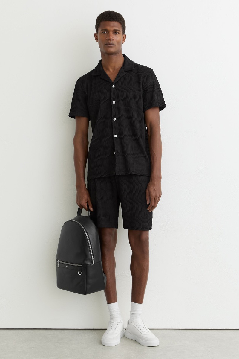 Man wearing a black textured short-sleeve camp collar shirt and matching black textured shorts with white crew socks and chunky white leather sneakers, holding a black leather backpack