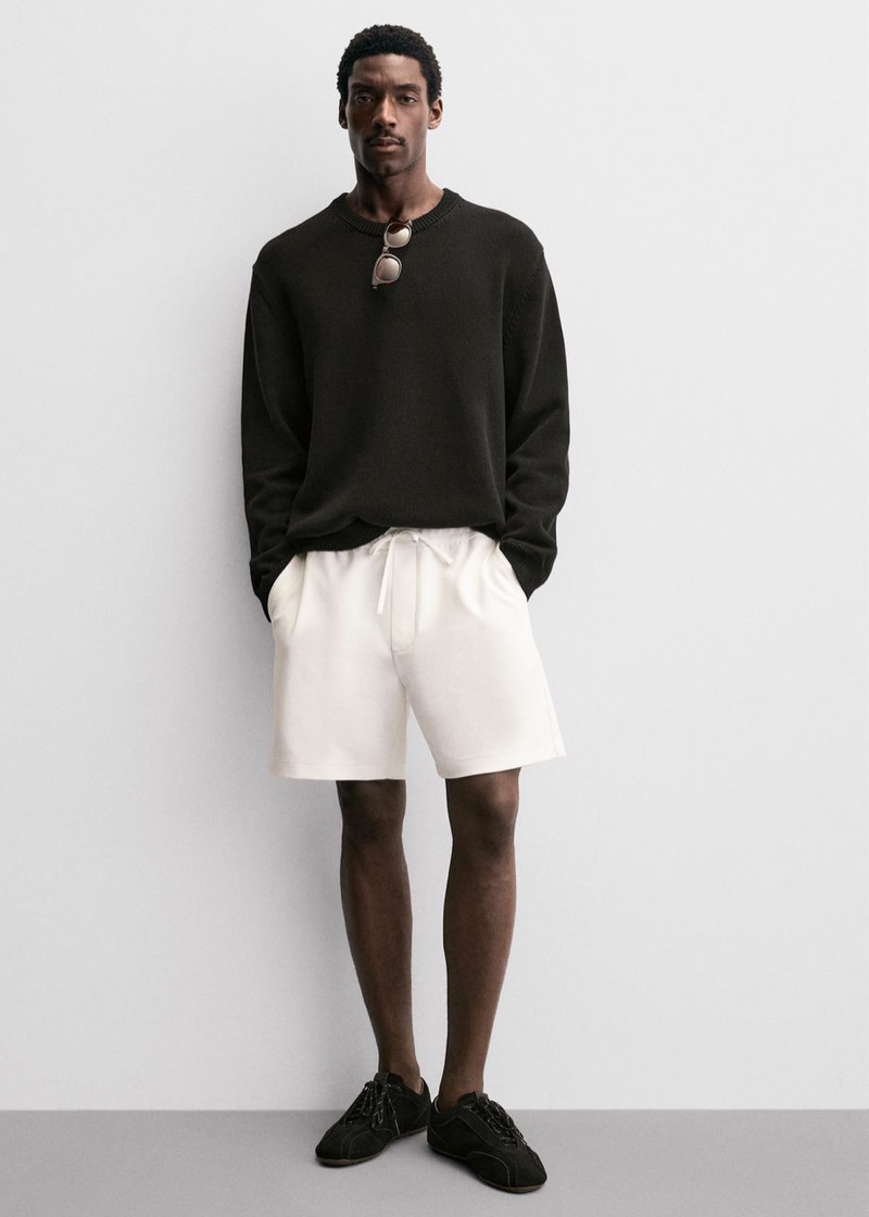 Man wearing white drawstring shorts with a black crewneck sweater and black sneakers