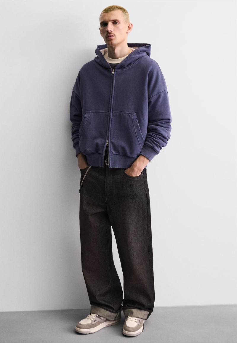 A young man with cropped blonde hair leans against a gray wall in a faded navy zip hoodie and baggy dark indigo jeans cuffed at the ankle, wearing off-white and taupe paneled low-top basketball sneakers.