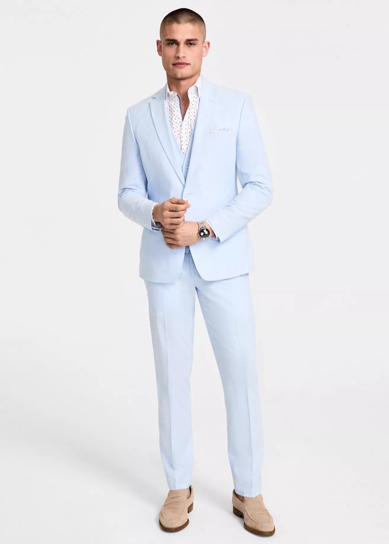 A man in a pale-blue three-piece linen-blend suit with notch lapels, a white pin-dot patterned camp-style shirt worn open-collar, a white pocket square, and tan suede penny loafers.