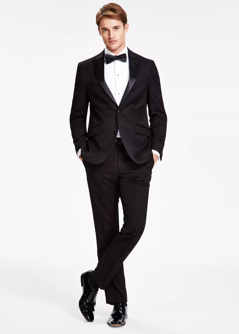 A man in a slim-fit black peak-lapel tuxedo with satin facings, a pleated white tuxedo shirt with black studs, a black silk bow tie, and patent leather black Oxford shoes.