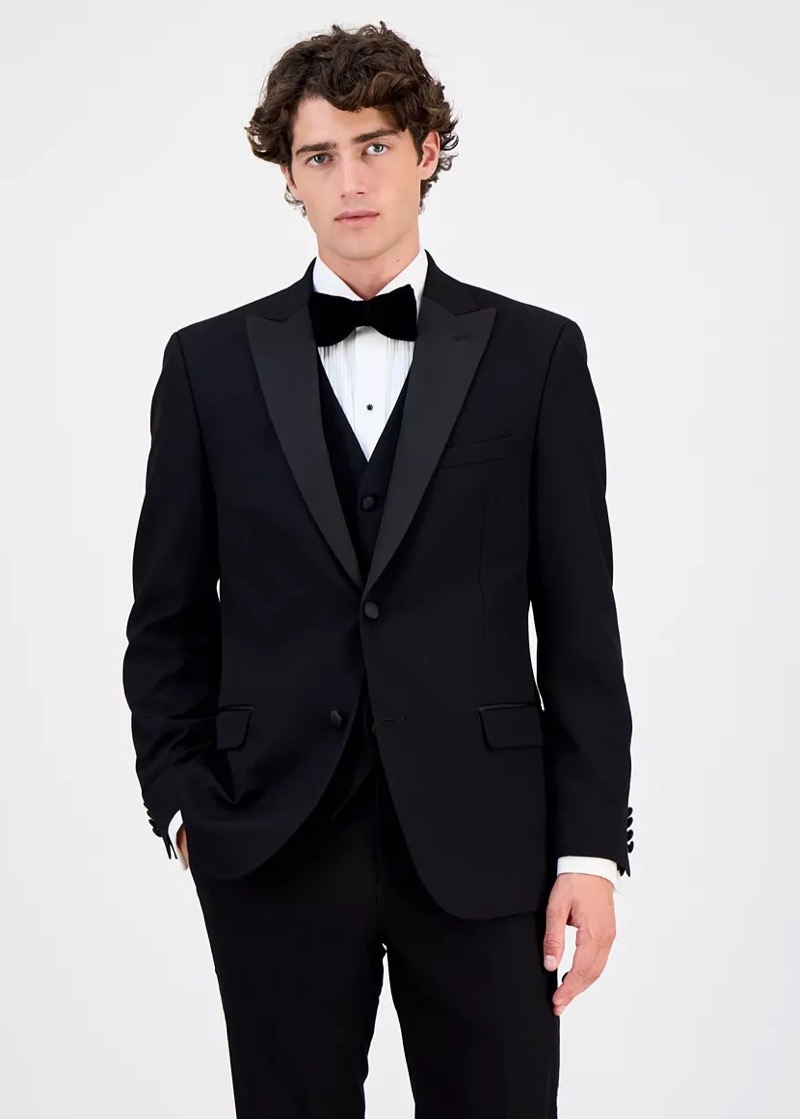 Front view of a man wearing a black peak-lapel tuxedo with satin facings, a matching black tuxedo waistcoat, a pleated white formal shirt with a single black stud, and a black silk bow tie.