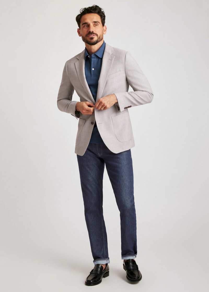 A bearded man buttons a light oatmeal-gray unstructured blazer with patch pockets over a teal-blue knit polo and dark indigo straight-leg jeans cuffed at the ankle, finished with black leather penny loafers.