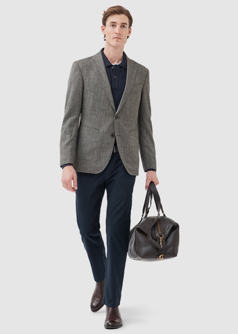 A man in a gray-and-cream herringbone unstructured blazer over a navy knit polo carries a dark brown leather weekender bag, paired with navy slim chinos and dark brown leather Chelsea boots.