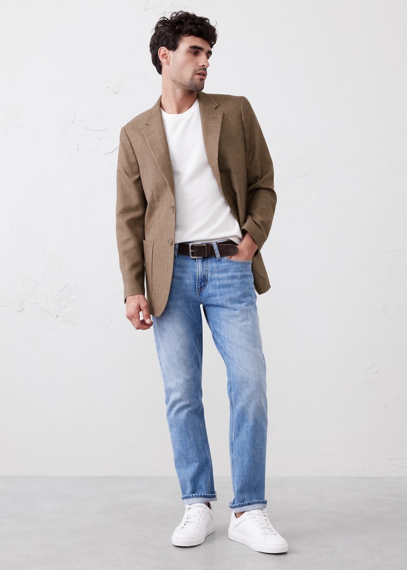 A man in a tan herringbone unstructured blazer over a plain white crew neck tee stands in light-wash straight-leg jeans cuffed at the hem above clean white leather low-top sneakers, belted with a dark brown leather strap.