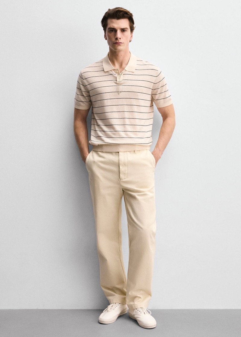 A man with combed-back hair stands against a pale wall in a cream knit polo with thin charcoal horizontal stripes and an open placket, tucked into wide-leg cream linen-cotton trousers and finished with off-white canvas sneakers.