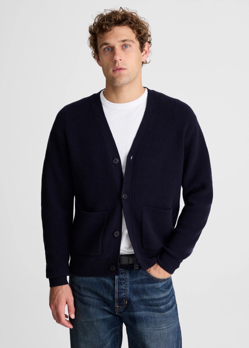A man with curly hair wears a chunky navy ribbed-knit V-neck cardigan with two front patch pockets and dark button closures over a white crew neck tee, paired with mid-wash jeans and a black belt.
