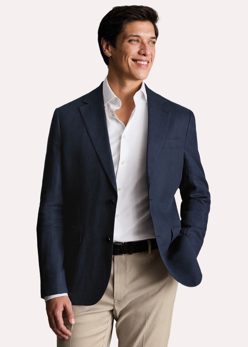 A smiling man wearing a navy linen-blend single-breasted blazer with notch lapels, an unbuttoned white oxford shirt, a brown leather belt, and stone-colored chino trousers.