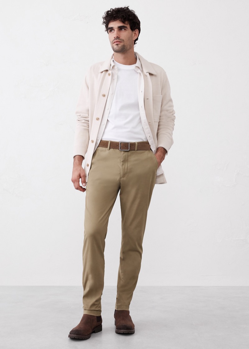 A man in a cream knit overshirt and white tee stands in tailored khaki flat-front chinos that taper through the leg to a clean break above dark brown suede Chelsea boots, cinched with a tan suede belt.