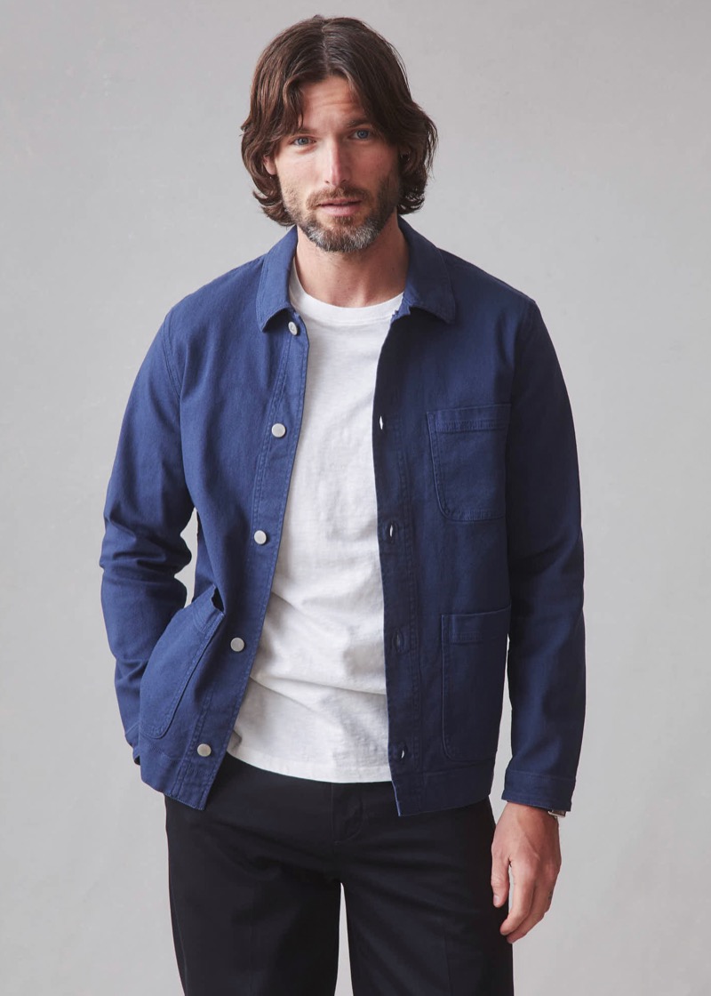 A bearded man wears an unbuttoned navy cotton chore coat with patch pockets, silver snap closures, and a sturdy collar over a heathered white crew neck tee and black trousers.