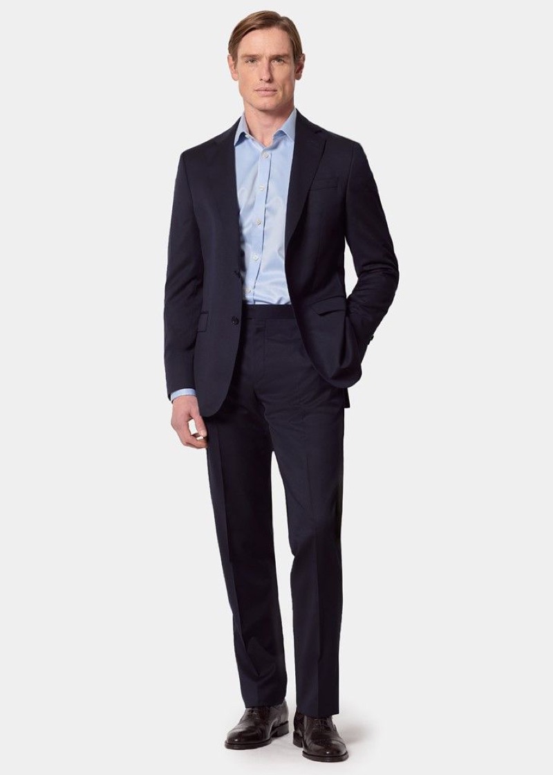A man in a dark navy slim-fit two-button suit with notch lapels and flat-front trousers, worn open-collar with a light blue dress shirt and dark brown leather brogues.