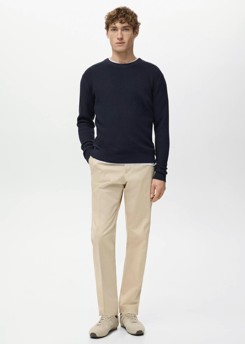 A young man stands in a textured navy ribbed crew neck sweater with a slim white tee just visible at the hem, tucked over flat-front cream chino trousers, finished with stone-gray suede runners.