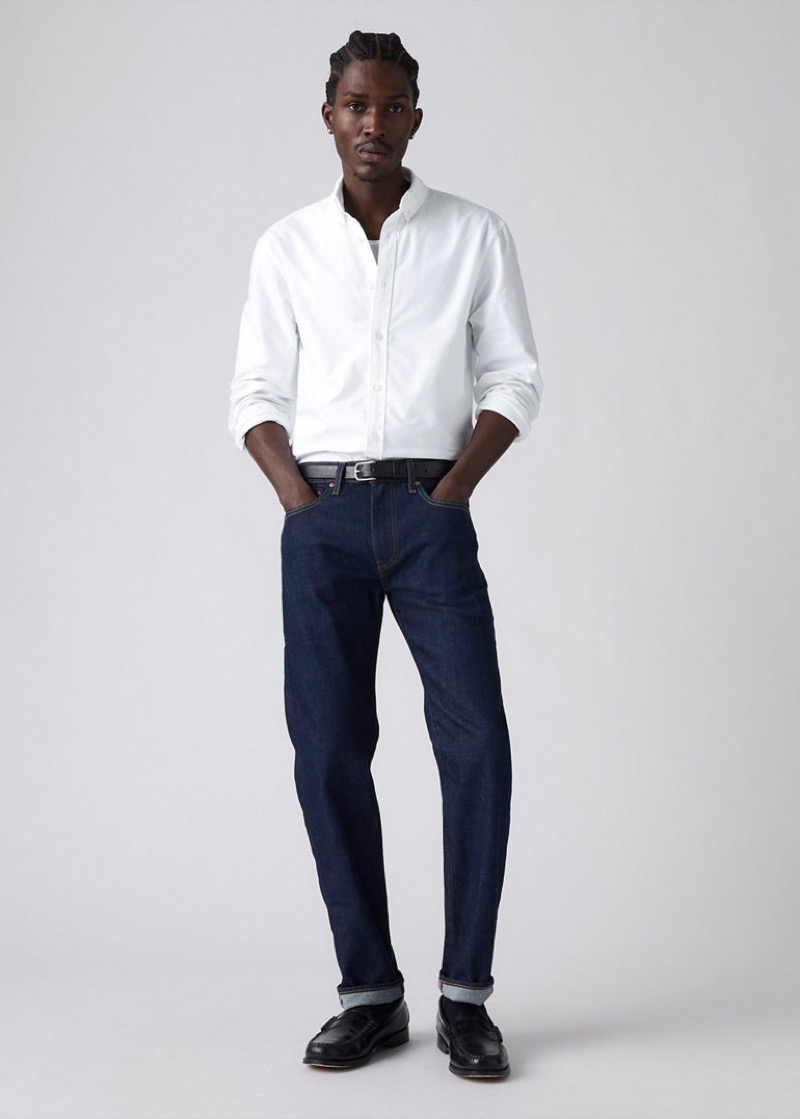 A man wears a crisp white oxford button-down shirt tucked into dark indigo rinse-wash tapered jeans with a black leather belt and silver buckle, rolled at the cuff over black leather penny loafers.