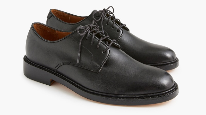 A pair of plain-toe black leather Derby shoes with open lacing, visible eyelet flaps sitting on top of the vamp, and tan leather lining.