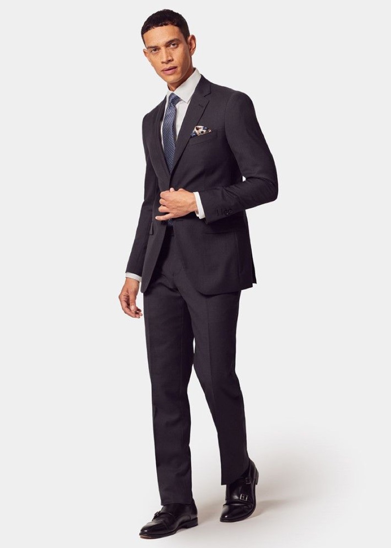 A man wearing a tailored charcoal worsted-wool two-button suit with notch lapels, a white dress shirt, a navy patterned silk tie, a patterned pocket square, and black double monk-strap shoes.
