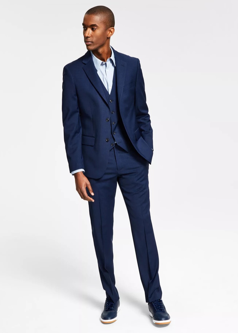 Full-length view of a man wearing a slim-fit royal-blue three-piece suit with matching waistcoat, a light blue dress shirt without a tie, and navy leather low-top sneakers.