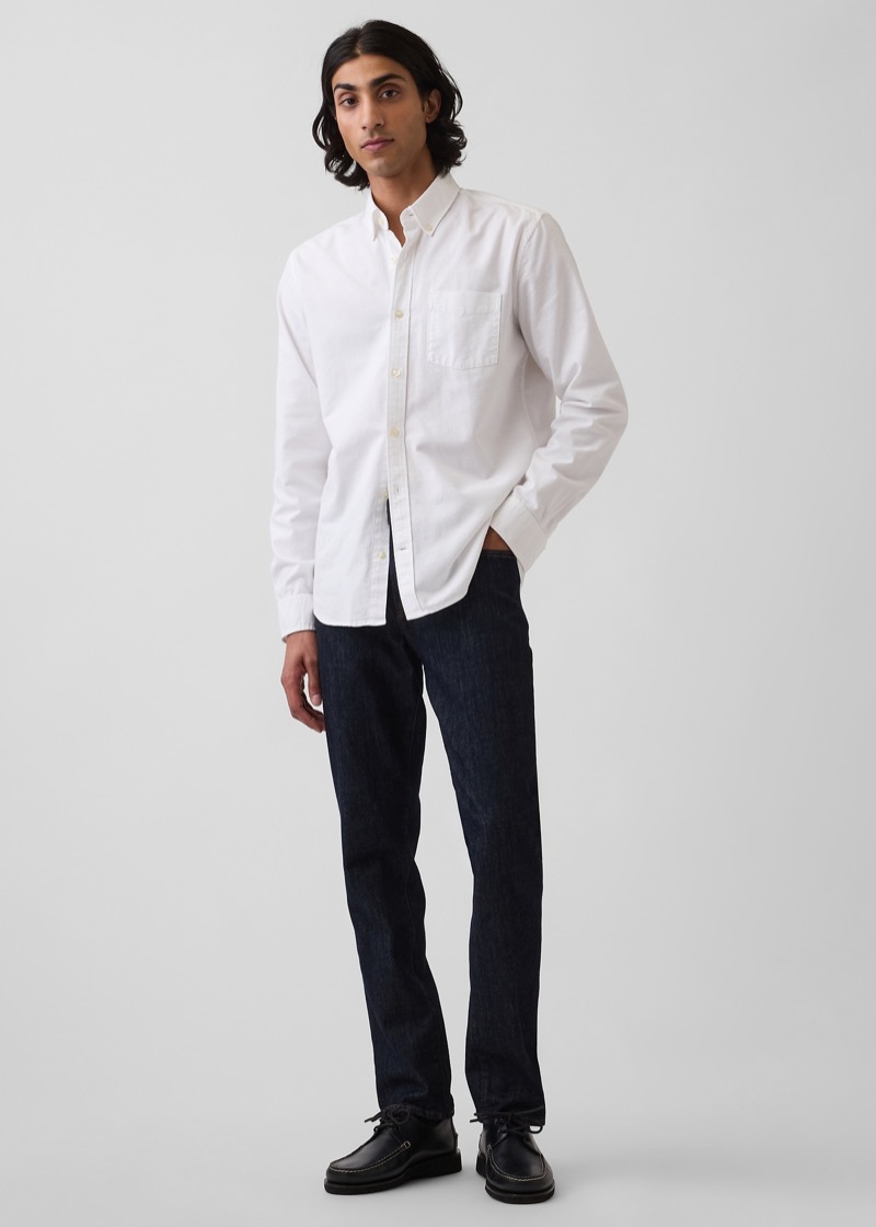 A man with long dark hair stands in a white oxford cloth button-down shirt with a chest patch pocket, half-tucked into dark indigo rinse-wash slim jeans, finished with black leather moc-toe shoes.