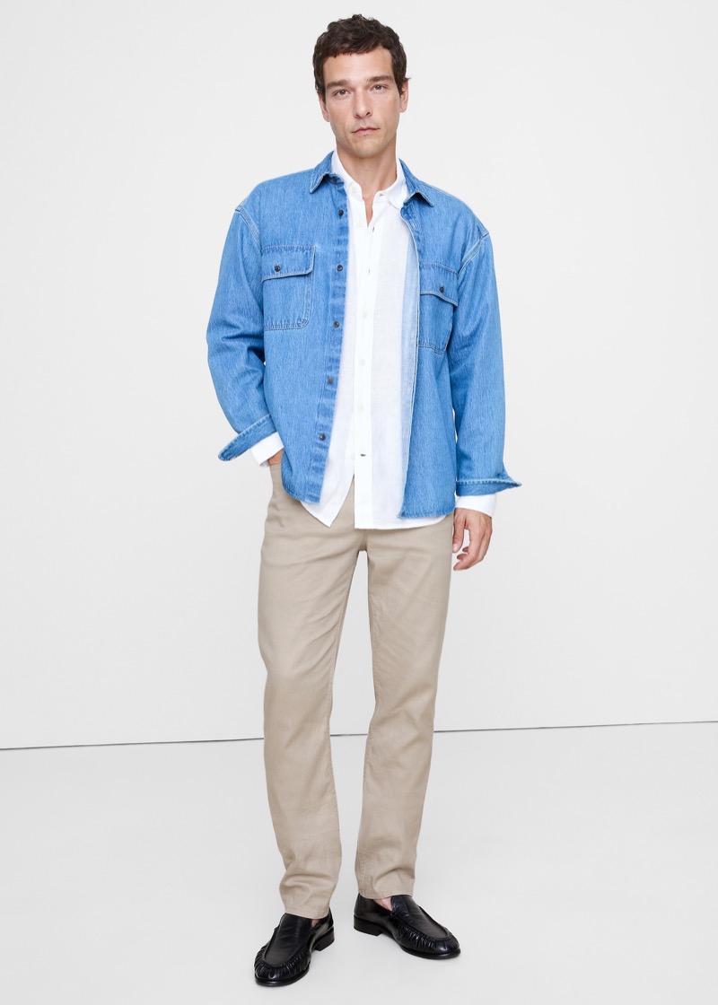 A man layers a mid-wash denim overshirt over a white linen button-down and stone linen-cotton trousers with a clean leg line, finished with black leather penny loafers.