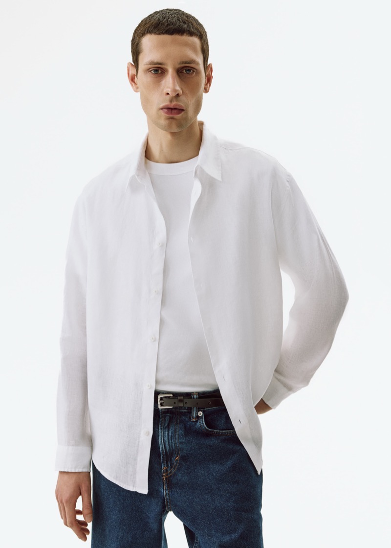A man with a buzz cut wears an unbuttoned white linen long-sleeve shirt with a relaxed silhouette over a white crew neck tee and mid-wash jeans, cinched with a slim dark brown leather belt.