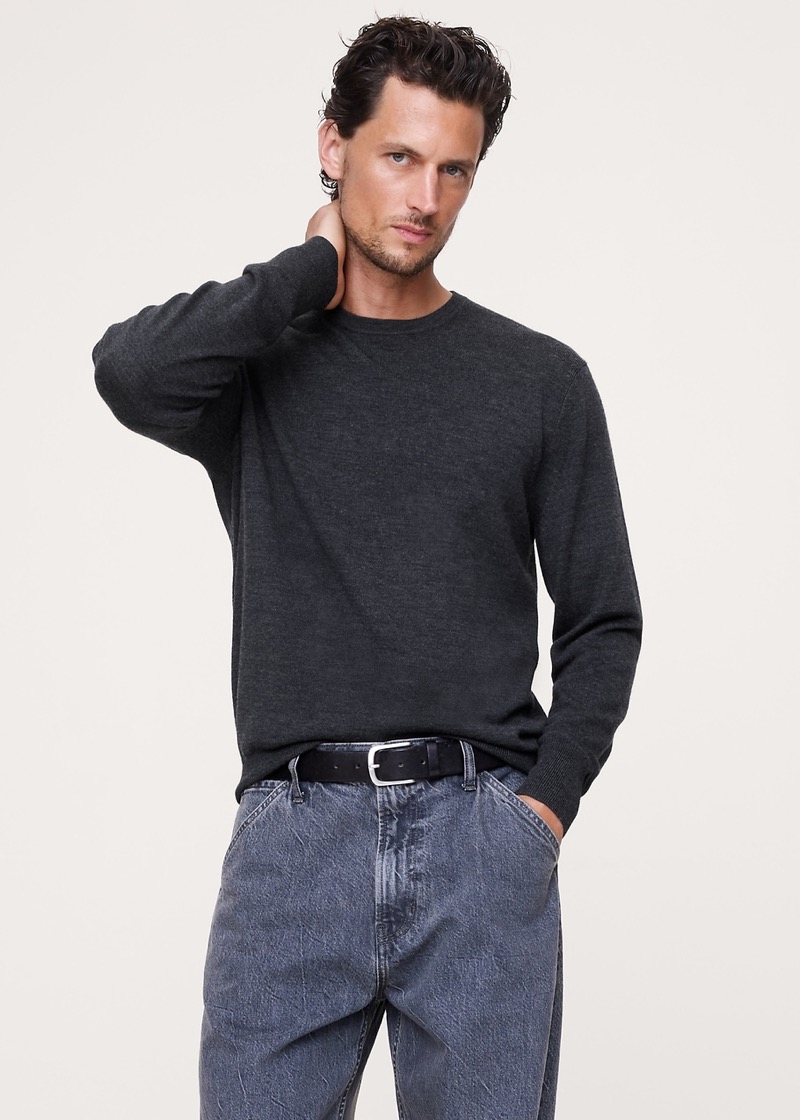 A man with wavy dark hair stands against a neutral backdrop in a charcoal-gray fine-gauge merino crew neck sweater with ribbed cuffs and hem, paired with a black leather belt and washed gray relaxed jeans.