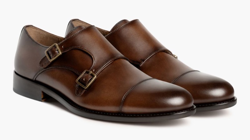 A pair of dark brown burnished leather double monk strap shoes with two antiqued silver buckles across the instep and a cap toe.