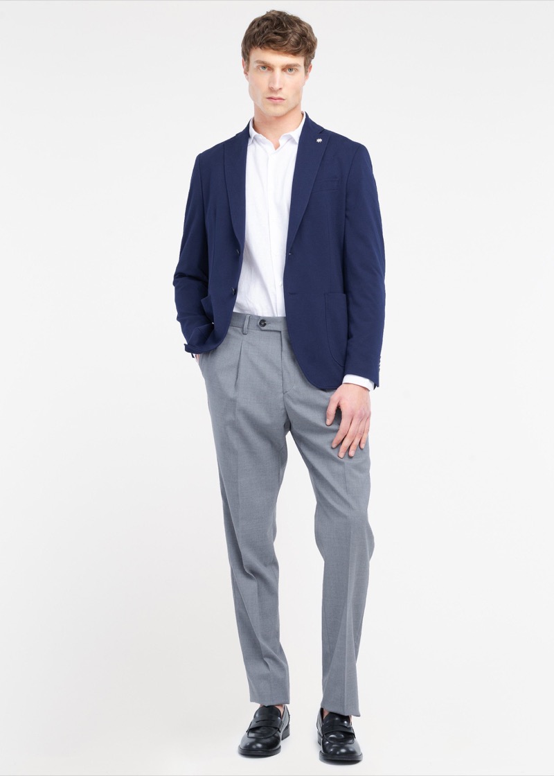 A man stands in front of a white backdrop wearing a navy unstructured two-button blazer with patch pockets and a small pin on the lapel, a white button-down shirt underneath, mid-gray pleated wool trousers tapering to a clean break, and black leather penny loafers.