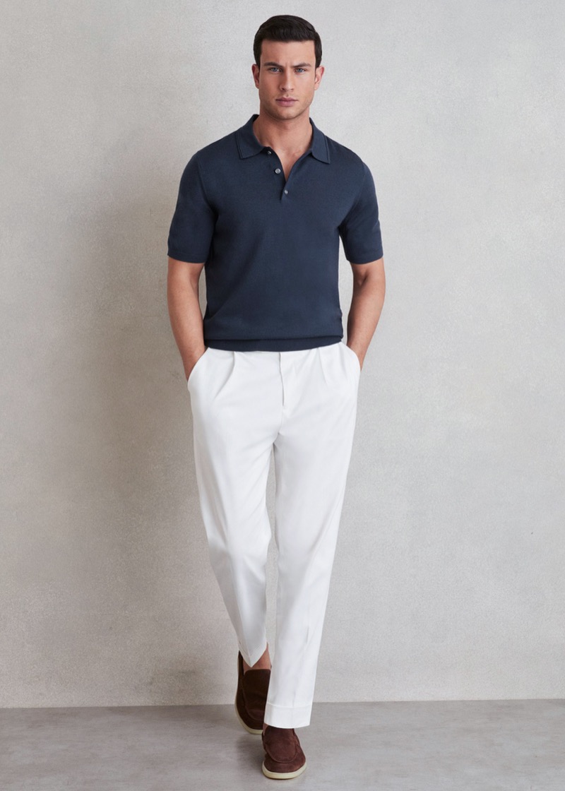 A man with short dark hair stands in a textured navy short-sleeve knit polo with an open Cuban collar, tucked into wide-leg ivory pleated trousers belted with a silver D-ring buckle, paired with dark brown suede loafers.