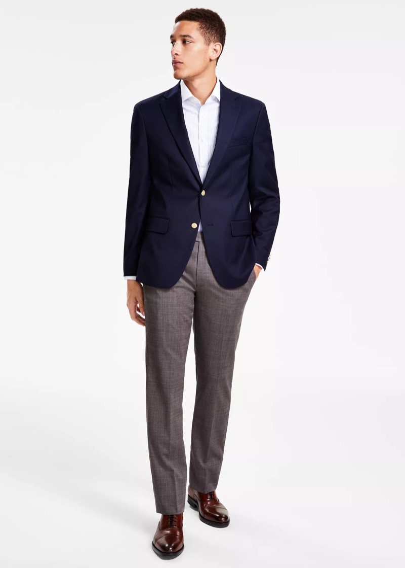 A man in a navy single-breasted blazer with gold buttons, worn with a fine-striped white dress shirt, taupe wool tailored trousers, and burgundy leather cap-toe Oxford shoes.