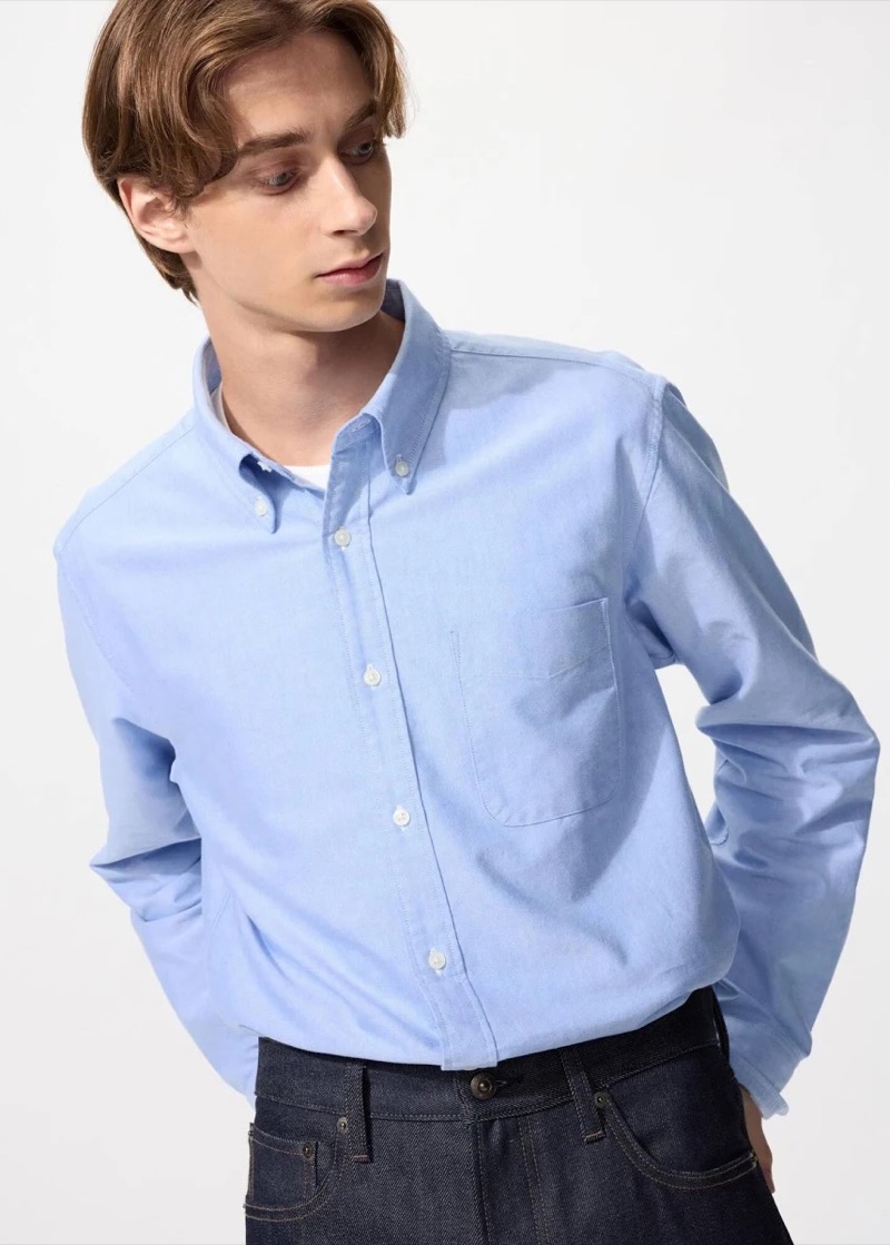 A young man with sweeping brown hair looks down while wearing a light blue oxford cloth button-down shirt with a soft button-down collar, a chest patch pocket, and white buttons, tucked into dark indigo jeans.