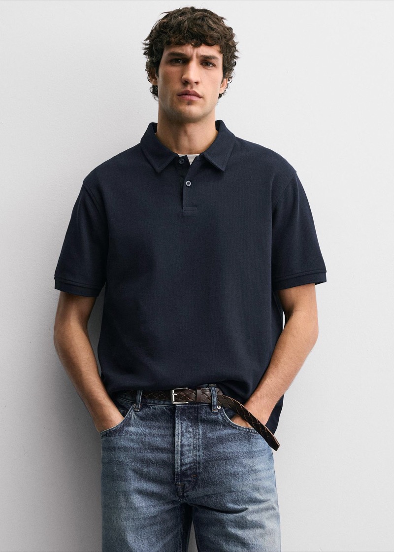 A young man with curly brown hair stands against a white wall in a relaxed-fit navy short-sleeve polo with a soft collar and a two-button placket, hands tucked into faded mid-blue jeans cinched with a braided dark brown leather belt.