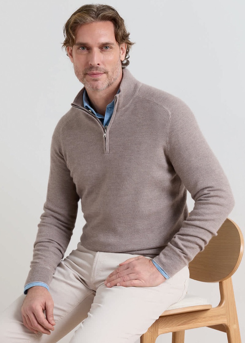 A man with curly brown hair and a beard sits on a wooden chair in a taupe merino quarter-zip sweater with raglan sleeves and a high zip collar, layered over a light blue chambray shirt with the collar peeking out, paired with cream chino trousers.
