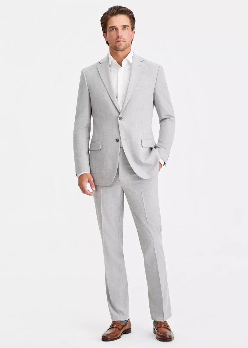 A man wearing a light gray slim-fit two-button suit with notch lapels, an open-collar white dress shirt, and dark brown leather penny loafers.
