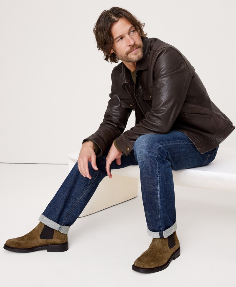 Man in a dark brown leather shirt jacket and rolled raw denim jeans sits on a low bench, wearing olive suede Chelsea boots with elastic side panels and a low stacked heel.