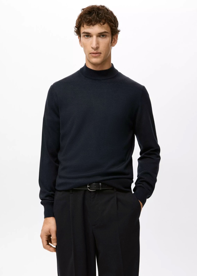 A young man stands against a pale wall in a fine-gauge navy mock-neck merino sweater with ribbed neck, cuffs, and hem, tucked into black pleated wool trousers with a slim black leather belt.